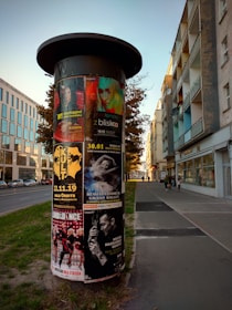 A cylindrical advertising column stands on a sidewalk, plastered with various colorful posters for concerts, dance performances, and theatrical events. The background features a bustling city street with modern buildings and a few people walking.