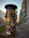 A cylindrical advertising column stands on a sidewalk, plastered with various colorful posters for concerts, dance performances, and theatrical events. The background features a bustling city street with modern buildings and a few people walking.