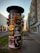 A cylindrical advertising column stands on a sidewalk, plastered with various colorful posters for concerts, dance performances, and theatrical events. The background features a bustling city street with modern buildings and a few people walking.