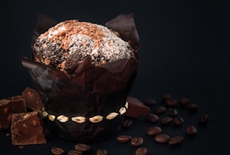 A richly chocolate-colored muffin sprinkled with powdered sugar and chocolate shavings is wrapped in dark paper sitting on a surface. Surrounding the muffin are scattered coffee beans and broken pieces of chocolate, creating a rustic and indulgent setting.