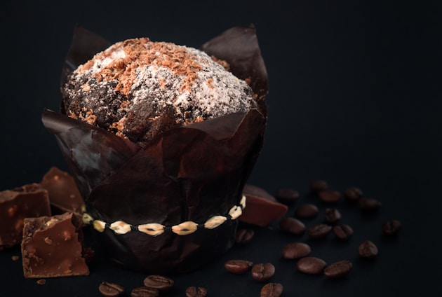 A richly chocolate-colored muffin sprinkled with powdered sugar and chocolate shavings is wrapped in dark paper sitting on a surface. Surrounding the muffin are scattered coffee beans and broken pieces of chocolate, creating a rustic and indulgent setting.
