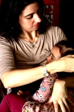 A woman with dark hair holds a small child in her arms. She is wearing a light-colored top, and the child is dressed in clothes with a floral pattern. Her expression is gentle and caring.