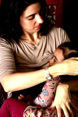 A woman with dark hair holds a small child in her arms. She is wearing a light-colored top, and the child is dressed in clothes with a floral pattern. Her expression is gentle and caring.