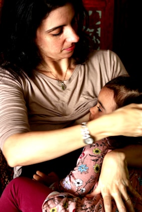 A woman with dark hair holds a small child in her arms. She is wearing a light-colored top, and the child is dressed in clothes with a floral pattern. Her expression is gentle and caring.