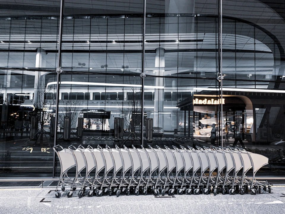 The image depicts a row of empty shopping carts lined up on a sidewalk, facing away from the camera and reflecting the lights from a nearby building. The perspective is from behind the carts, giving an impression that they are standing in front of a large glass window or entrance to a store. In the background, there is a sign for "MAD" visible through the window.
