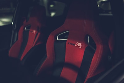 A close-up of two sporty car seats with bright red fabric and black accents. The seats have the branding 'Type R' embroidered on them and feature a distinctive ergonomic design.