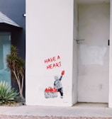 A stencil art piece on a white wall depicts a child holding up a red heart. Next to the child is a basket filled with more red hearts. The words 'HAVE A HEART' are written above in bold red letters. There are plants nearby on the left, adding some greenery beside the artwork. The setting appears to be an urban environment.
