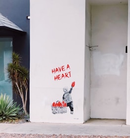 A stencil art piece on a white wall depicts a child holding up a red heart. Next to the child is a basket filled with more red hearts. The words 'HAVE A HEART' are written above in bold red letters. There are plants nearby on the left, adding some greenery beside the artwork. The setting appears to be an urban environment.