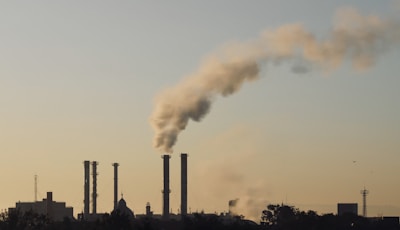 Several industrial chimneys emit thick smoke into the sky, silhouetted against a pale and hazy background. The skyline includes buildings and structures, with sparse vegetation in the foreground.