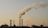 Several industrial chimneys emit thick smoke into the sky, silhouetted against a pale and hazy background. The skyline includes buildings and structures, with sparse vegetation in the foreground.
