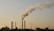 Several industrial chimneys emit thick smoke into the sky, silhouetted against a pale and hazy background. The skyline includes buildings and structures, with sparse vegetation in the foreground.