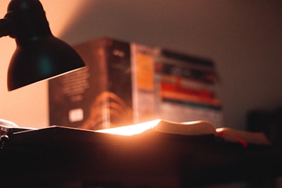 A warm glowing table lamp casting soft light over a wooden desk with a stack of books.