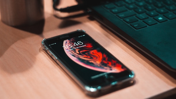A close-up of a sleek smartphone resting on a wooden desk beside a laptop and a cup of coffee.