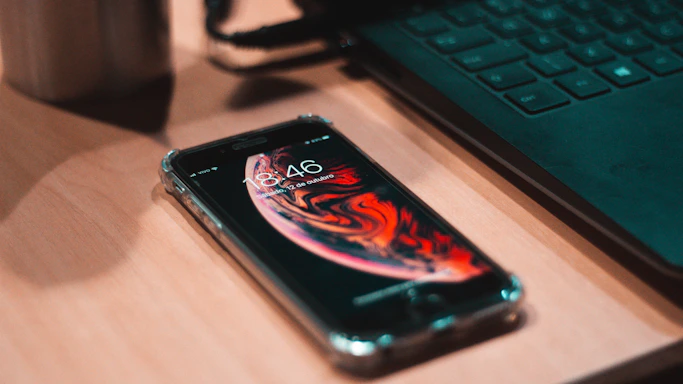 A modern smartphone lying on a sleek desk, next to a laptop and a blue coffee mug.