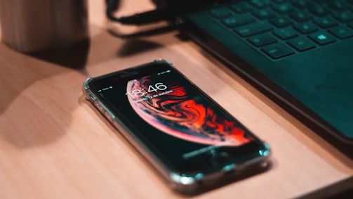 A sleek smartphone resting on a wooden desk beside a laptop and a cup of coffee.