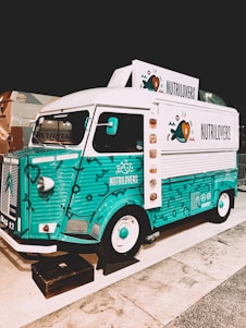 A colorful vintage food truck with a teal and white design, featuring the brand name 'NUTRILOVERS' and decorative graphics of hearts and fish. The front part of the truck showcases a distinctive logo, and the vehicle is parked on a paved area next to another truck.