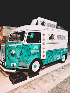 A colorful vintage food truck with a teal and white design, featuring the brand name 'NUTRILOVERS' and decorative graphics of hearts and fish. The front part of the truck showcases a distinctive logo, and the vehicle is parked on a paved area next to another truck.