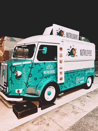 A colorful vintage food truck with a teal and white design, featuring the brand name 'NUTRILOVERS' and decorative graphics of hearts and fish. The front part of the truck showcases a distinctive logo, and the vehicle is parked on a paved area next to another truck.