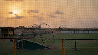 A baseball field is seen with a batting cage in the foreground, under a cloudy sky during sunset. The scene is enveloped in a warm glow from the setting sun. Few people are visible on the field, possibly practicing.