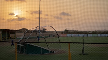 A baseball field is seen with a batting cage in the foreground, under a cloudy sky during sunset. The scene is enveloped in a warm glow from the setting sun. Few people are visible on the field, possibly practicing.
