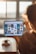 A person holding a tablet with a photo editing app open on the screen, featuring various images and templates. The setting appears to be a bright room, possibly a home or office, with sunlight streaming through large windows in the background.