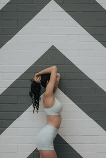 A person stands in profile with arms raised over the head, wearing light-colored athletic clothing. The background features a brick wall painted with large geometric chevrons in shades of white and gray.