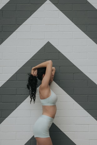 A person stands in profile with arms raised over the head, wearing light-colored athletic clothing. The background features a brick wall painted with large geometric chevrons in shades of white and gray.