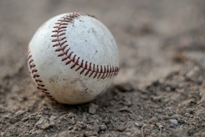 A worn-out baseball resting on rough dirt, displaying visible stitching and scuff marks, suggesting frequent use. The close-up perspective highlights texture and detail.