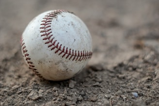 A worn-out baseball resting on rough dirt, displaying visible stitching and scuff marks, suggesting frequent use. The close-up perspective highlights texture and detail.