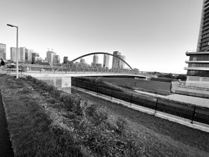 A modern arched bridge spans over a railway or canal, set against a backdrop of high-rise buildings. The scene features a grassy foreground with a path and streetlamp on the left side. The architecture presents a mix of contemporary urban elements, with construction cranes visible in the distance.