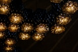 A series of hexagonal light fixtures hanging from a ceiling, each emitting a warm, yellowish light. The fixtures are made of numerous small, reflective elements, creating a textured, almost mosaic-like appearance. The background shows a dark, ribbed surface, possibly wooden panels, enhancing the contrast with the glowing lights.