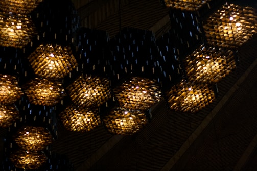 A series of hexagonal light fixtures hanging from a ceiling, each emitting a warm, yellowish light. The fixtures are made of numerous small, reflective elements, creating a textured, almost mosaic-like appearance. The background shows a dark, ribbed surface, possibly wooden panels, enhancing the contrast with the glowing lights.