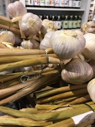 A close-up view of several heads of garlic bound together with wire. The garlic is displayed in a market or store setting, with shelves of bottles in the background out of focus. The stalks of the garlic are visible and appear dried.