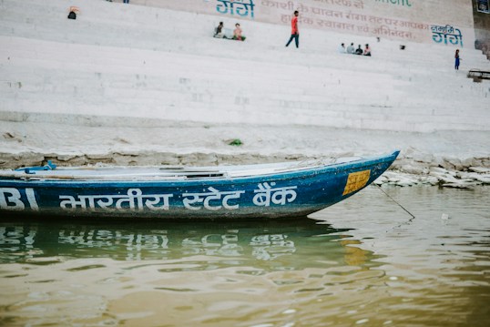 A wooden boat with Hindi script painted on its side floats on a river. The background features stone steps leading up to a wall with more Hindi text. People are sitting or walking on the steps, suggesting a lively environment.