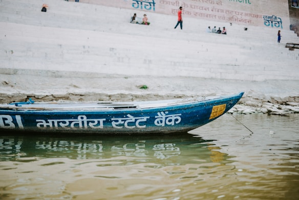 A wooden boat with Hindi script painted on its side floats on a river. The background features stone steps leading up to a wall with more Hindi text. People are sitting or walking on the steps, suggesting a lively environment.
