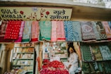 A vibrant market stall displaying an array of colorful textiles hanging on the front wall. A man sits amidst baskets filled with red rose petals. Shelved books and other items are visible in the background, and the store is adorned with a sign in multiple languages.