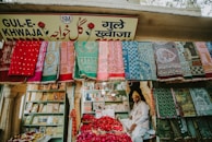 A vibrant market stall displaying an array of colorful textiles hanging on the front wall. A man sits amidst baskets filled with red rose petals. Shelved books and other items are visible in the background, and the store is adorned with a sign in multiple languages.