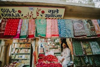 A vibrant market stall displaying an array of colorful textiles hanging on the front wall. A man sits amidst baskets filled with red rose petals. Shelved books and other items are visible in the background, and the store is adorned with a sign in multiple languages.