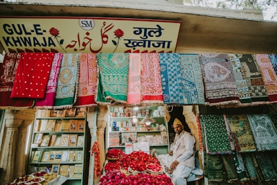 A vibrant market stall displaying an array of colorful textiles hanging on the front wall. A man sits amidst baskets filled with red rose petals. Shelved books and other items are visible in the background, and the store is adorned with a sign in multiple languages.