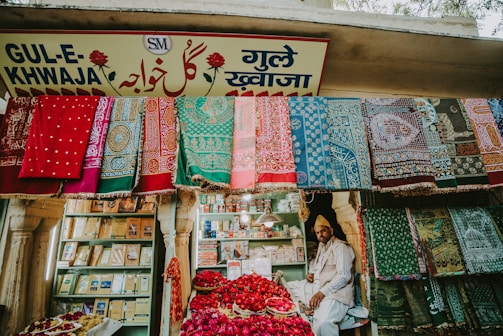 A vibrant market stall displaying an array of colorful textiles hanging on the front wall. A man sits amidst baskets filled with red rose petals. Shelved books and other items are visible in the background, and the store is adorned with a sign in multiple languages.