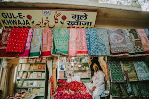 A vibrant market stall displaying an array of colorful textiles hanging on the front wall. A man sits amidst baskets filled with red rose petals. Shelved books and other items are visible in the background, and the store is adorned with a sign in multiple languages.