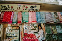 A vibrant market stall displaying an array of colorful textiles hanging on the front wall. A man sits amidst baskets filled with red rose petals. Shelved books and other items are visible in the background, and the store is adorned with a sign in multiple languages.