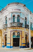 A vibrant, ornately designed corner building with intricate decorative elements, featuring a white and yellow façade accented with blue trim. The structure contains multiple arched windows and doors, with the name 'M RASCON' displayed prominently near the top. A sign for 'Funerales Puebla' is visible above one entrance, suggesting a funeral service establishment.