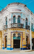 A vibrant, ornately designed corner building with intricate decorative elements, featuring a white and yellow façade accented with blue trim. The structure contains multiple arched windows and doors, with the name 'M RASCON' displayed prominently near the top. A sign for 'Funerales Puebla' is visible above one entrance, suggesting a funeral service establishment.