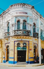 A vibrant, ornately designed corner building with intricate decorative elements, featuring a white and yellow façade accented with blue trim. The structure contains multiple arched windows and doors, with the name 'M RASCON' displayed prominently near the top. A sign for 'Funerales Puebla' is visible above one entrance, suggesting a funeral service establishment.