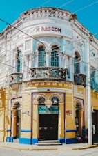 A vibrant, ornately designed corner building with intricate decorative elements, featuring a white and yellow façade accented with blue trim. The structure contains multiple arched windows and doors, with the name 'M RASCON' displayed prominently near the top. A sign for 'Funerales Puebla' is visible above one entrance, suggesting a funeral service establishment.