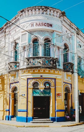 A vibrant, ornately designed corner building with intricate decorative elements, featuring a white and yellow façade accented with blue trim. The structure contains multiple arched windows and doors, with the name 'M RASCON' displayed prominently near the top. A sign for 'Funerales Puebla' is visible above one entrance, suggesting a funeral service establishment.