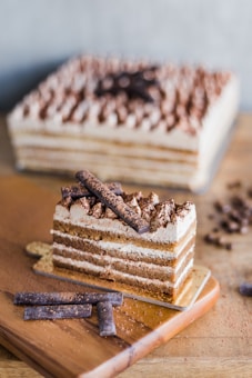 A slice of layered tiramisu cake sits elegantly on a wooden board, topped with chocolate curls and a dusting of cocoa powder. In the background, an entire tiramisu is visible, displaying its creamy layers and decorative top. The cake's texture appears rich and moist, with alternating layers of coffee-soaked cake and creamy filling.