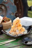 A dish featuring a pile of sushi rolls topped with shredded crab and creamy orange sauce, served on a black ceramic plate. Nearby, there is a bamboo steamer with pickled vegetables, a set of chopsticks, and a small dish of soy sauce. The rustic background includes a blue and rust-colored metal wheel, enhancing the outdoor dining setup.