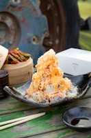 A dish featuring a pile of sushi rolls topped with shredded crab and creamy orange sauce, served on a black ceramic plate. Nearby, there is a bamboo steamer with pickled vegetables, a set of chopsticks, and a small dish of soy sauce. The rustic background includes a blue and rust-colored metal wheel, enhancing the outdoor dining setup.